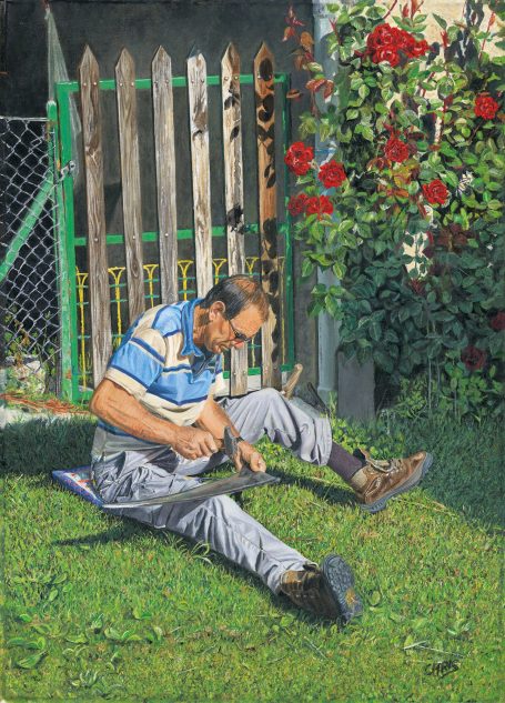 Un homme assis sur l'herbe, travaillant sur un objet, devant une clôture en bois et des fleurs.